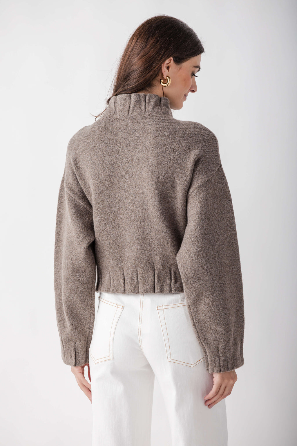 Beige knit jacket with pleated ruffle hem detail at waist, worn with white jeans, shown from back.