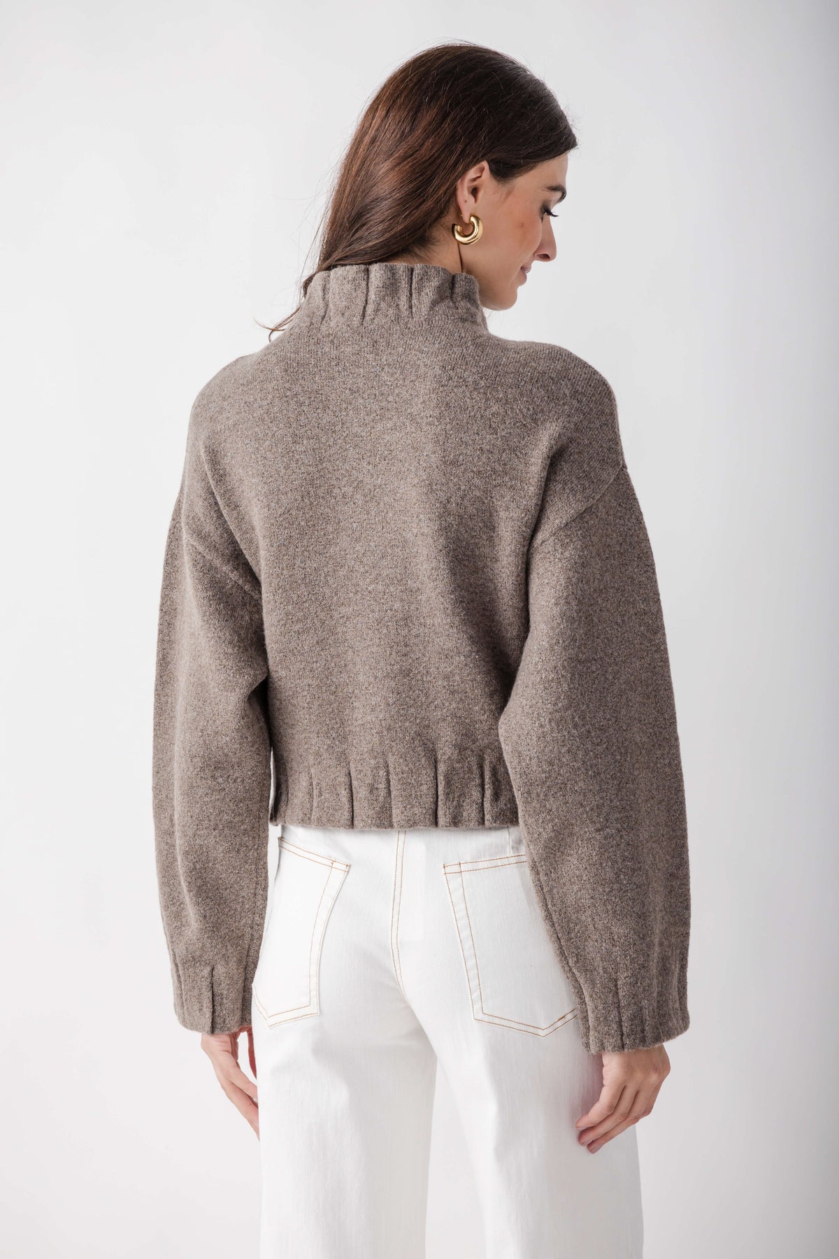 Beige knit jacket with pleated ruffle hem detail at waist, worn with white jeans, shown from back.
