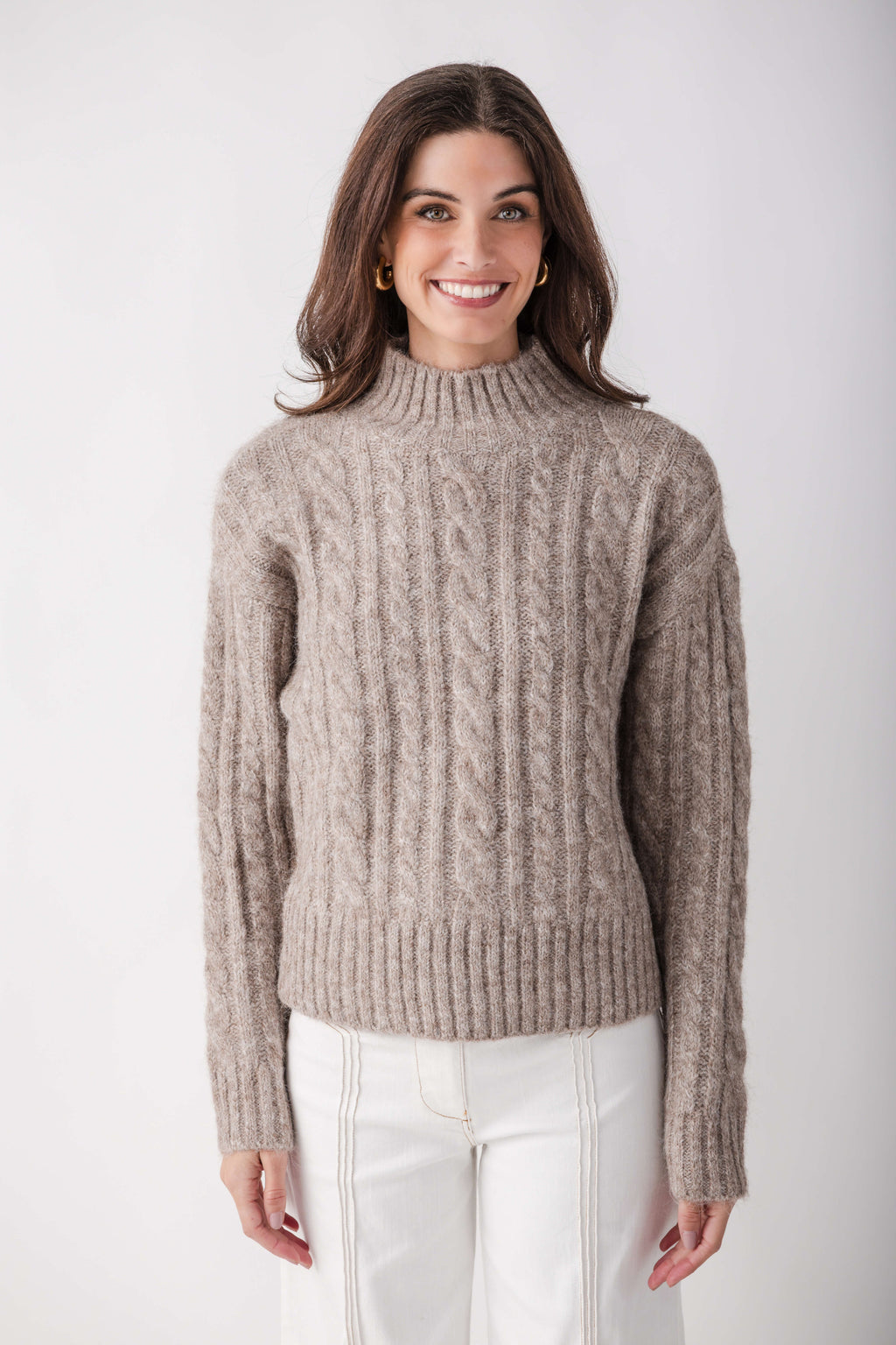 Beige cable knit turtleneck sweater with ribbed cuffs and hem worn with white pants.