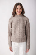 Beige cable knit turtleneck sweater with ribbed cuffs and hem worn with white pants.