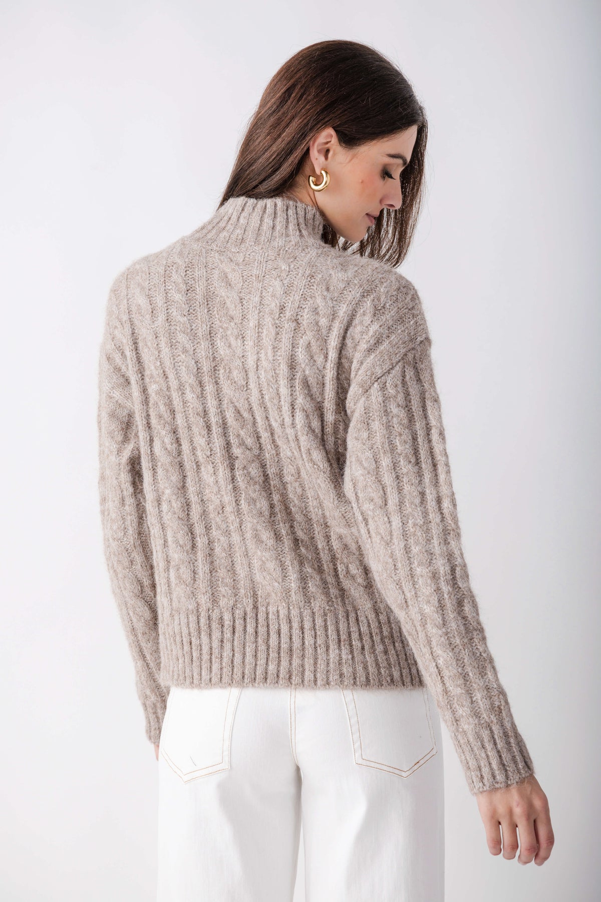 Beige cable knit turtleneck sweater with ribbed hem and cuffs shown from back view on model wearing white jeans.