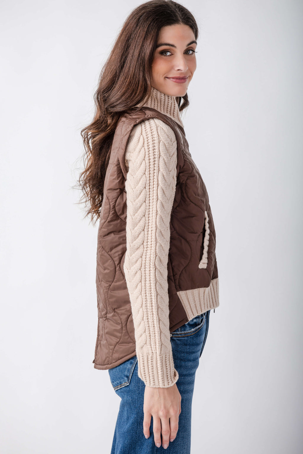 Brown quilted jacket with cream cable-knit sleeves and turtleneck collar, worn over blue jeans.