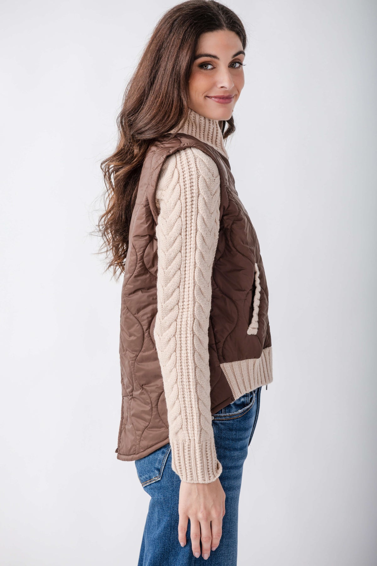 Brown quilted jacket with cream cable-knit sleeves and turtleneck collar, worn over blue jeans.