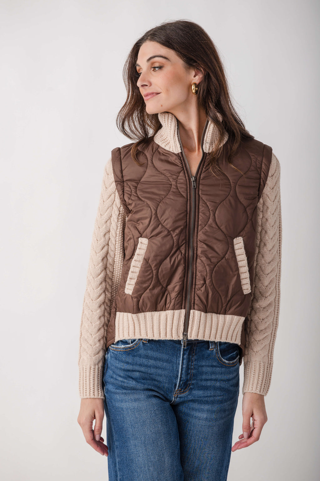 Brown quilted vest with cream knit sleeves, ribbed trim, and full-length zipper worn over blue jeans.