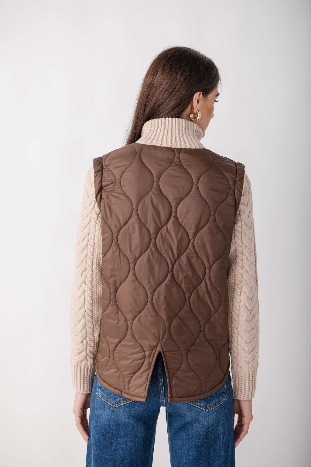 Woman wearing a mixed media jacket with a quilted brown vest over cream cable-knit sleeves, paired with blue jeans.
