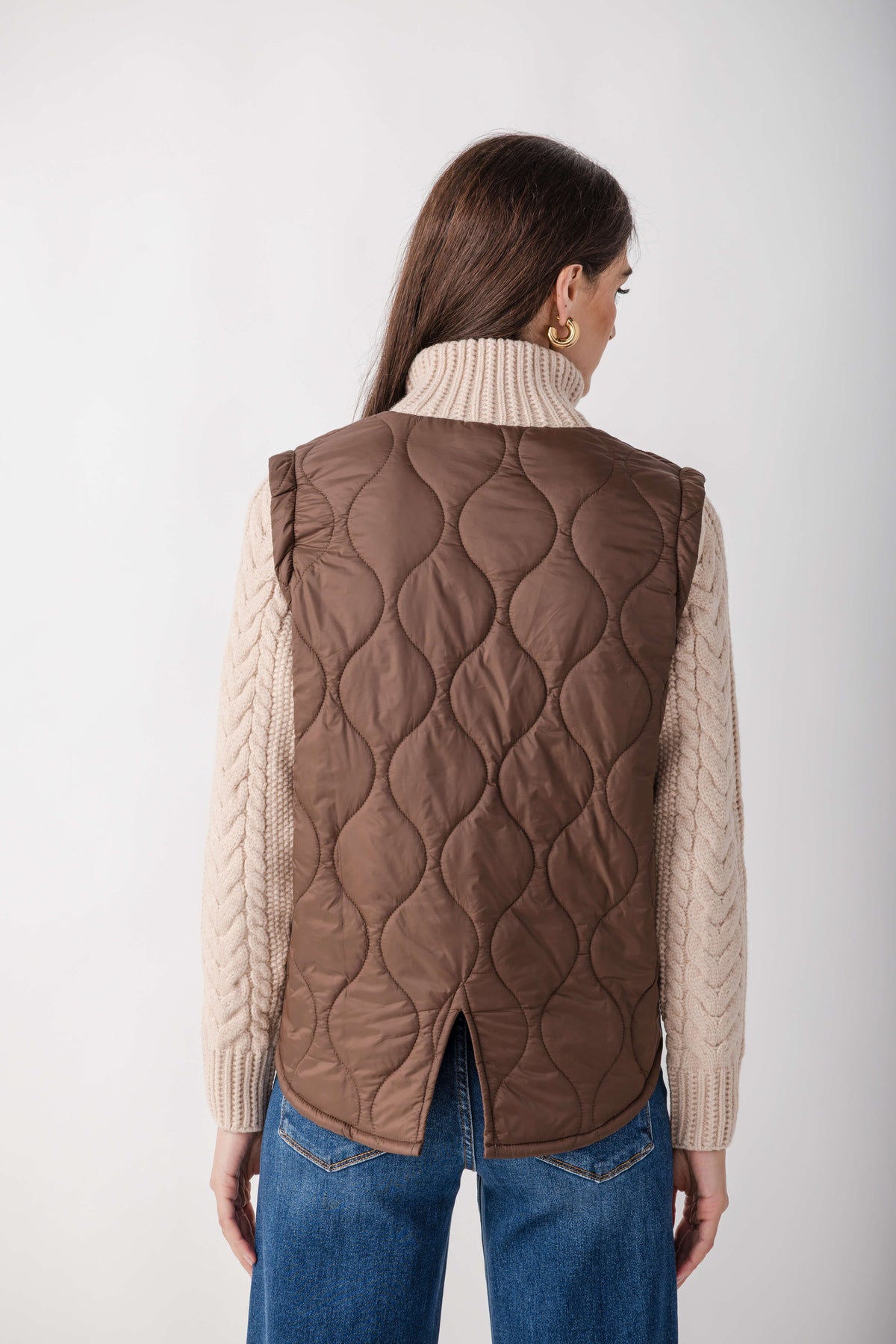 Woman wearing a mixed media jacket with a quilted brown vest over cream cable-knit sleeves, paired with blue jeans.
