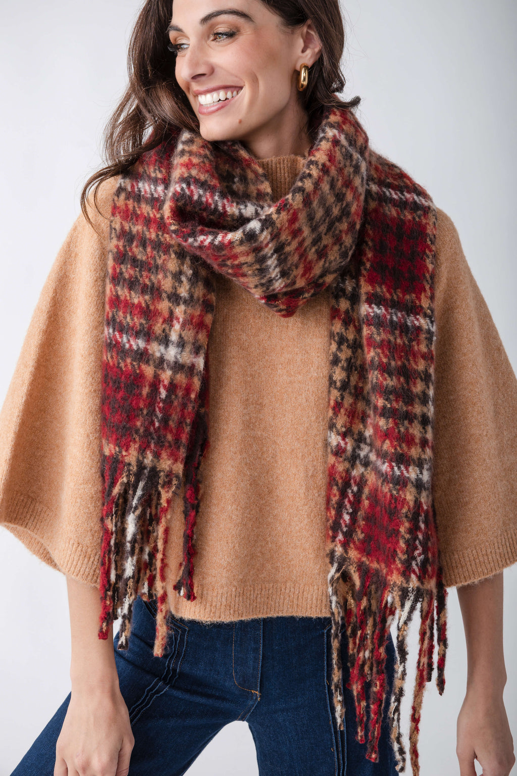 Red, cream, and black houndstooth scarf with fringe trim draped around woman's shoulders over tan sweater.