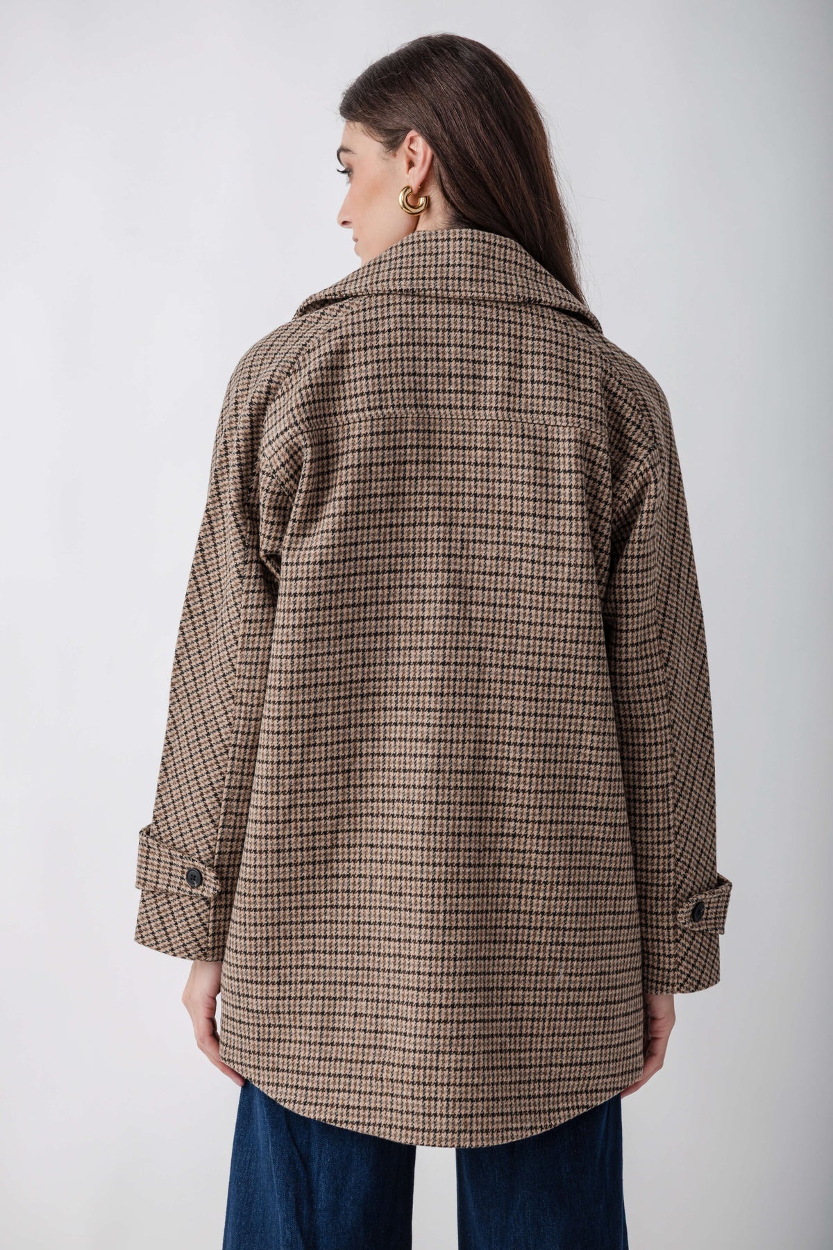 Woman wearing a tan and brown houndstooth pea jacket with a collared neckline and rolled sleeves, shown from the back.