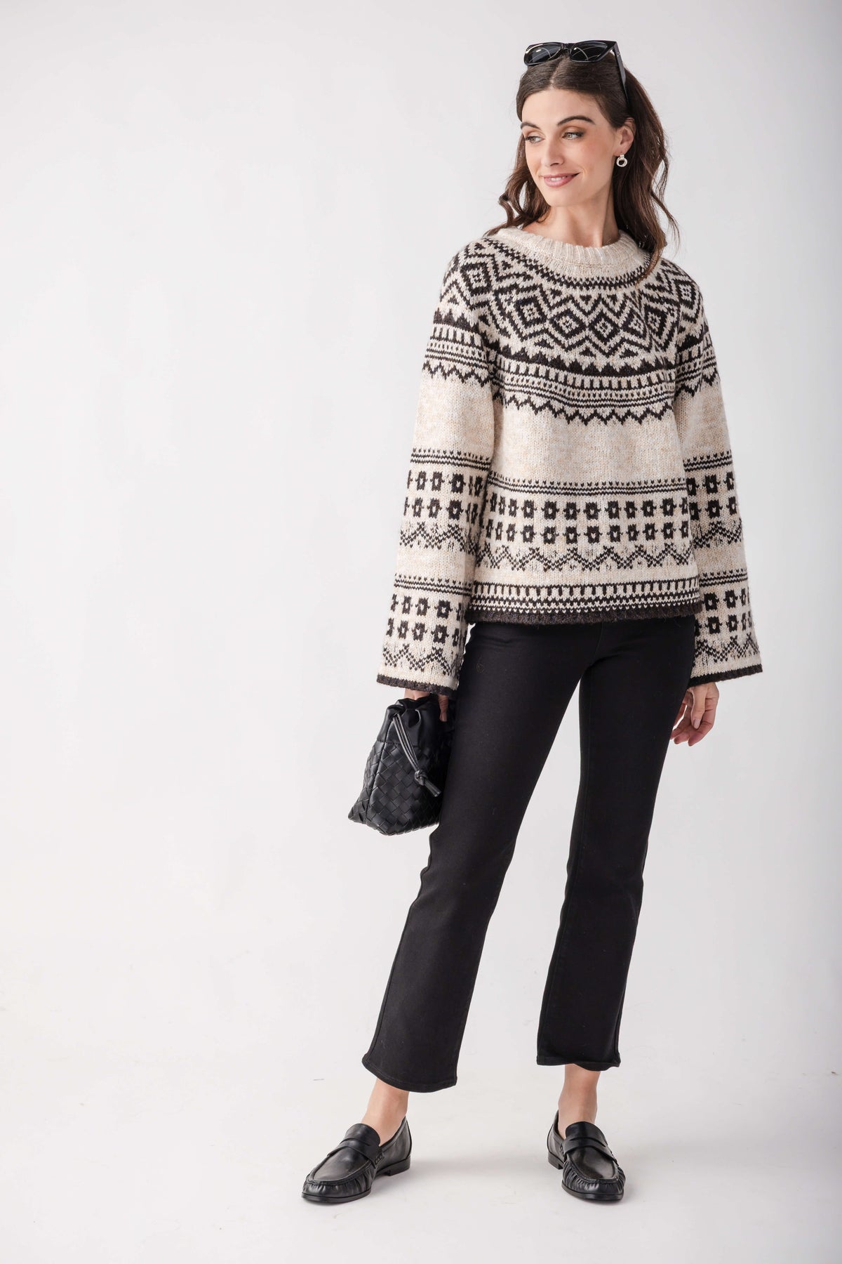 A woman modeling a cream and black patterned knit sweater with bell sleeves, paired with black pants and black loafers, against a light gray studio background.