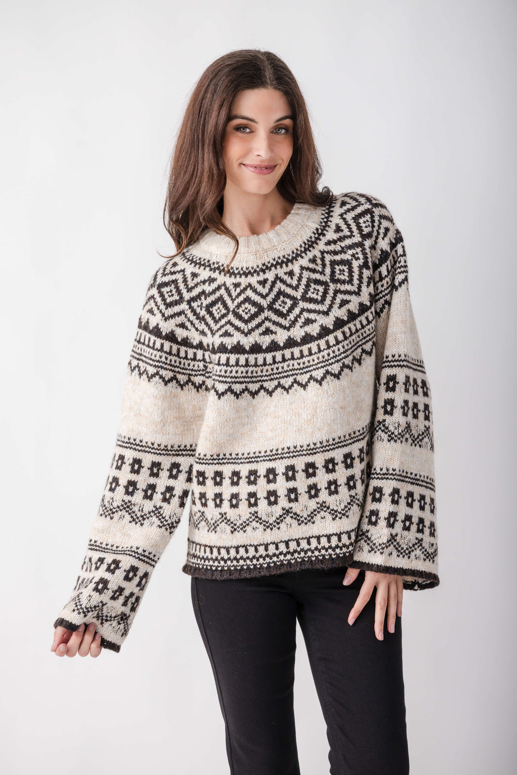 A woman wears a cream knit sweater with black Nordic geometric patterns, wide sleeves, and a crew neckline, paired with black pants.