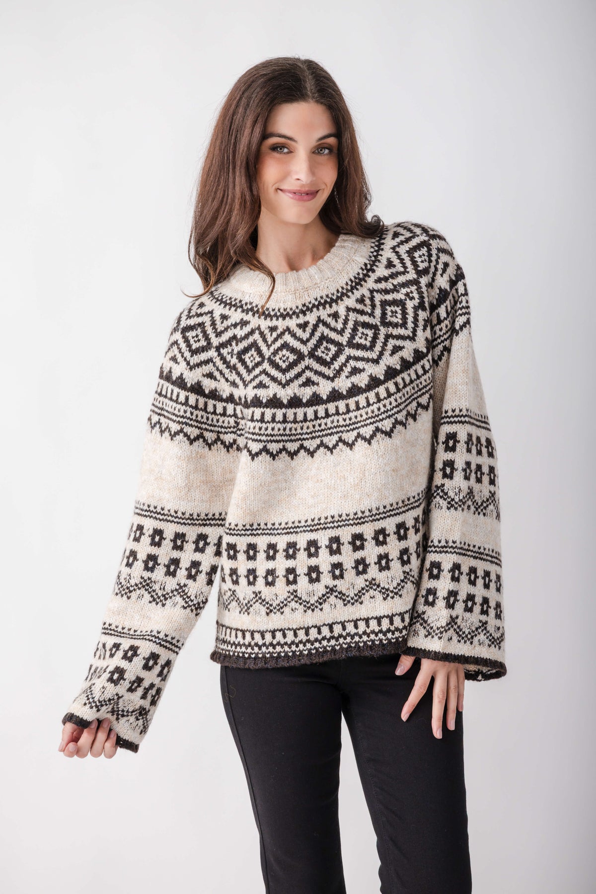 A woman wears a cream knit sweater with black Nordic geometric patterns, wide sleeves, and a crew neckline, paired with black pants.