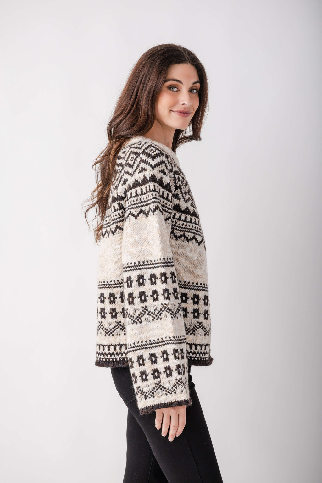 A woman with long brown hair wears a cream knit sweater with black Nordic patterns and black pants, glancing over her shoulder.