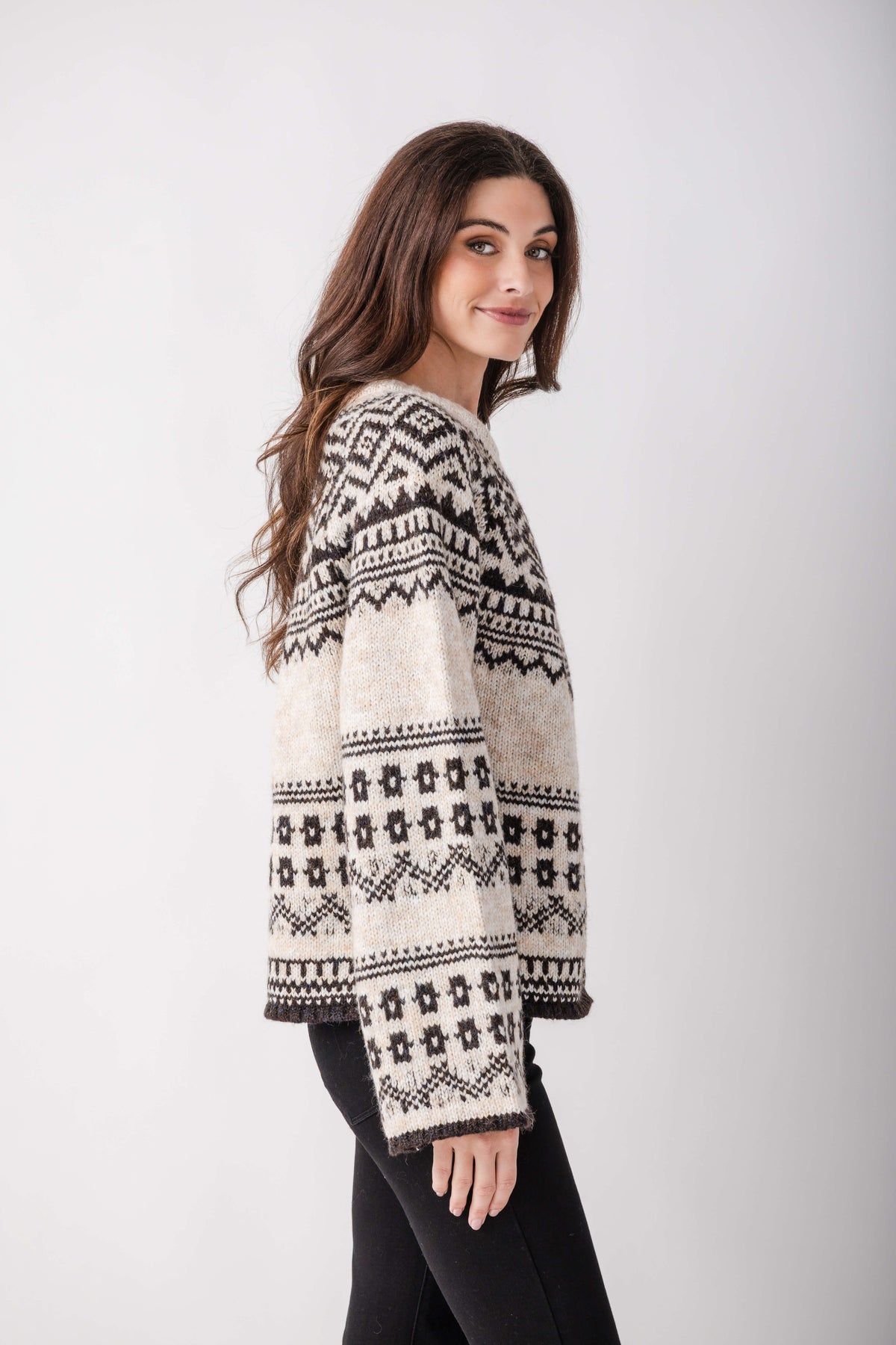 A woman with long brown hair wears a cream knit sweater with black Nordic patterns and black pants, glancing over her shoulder.