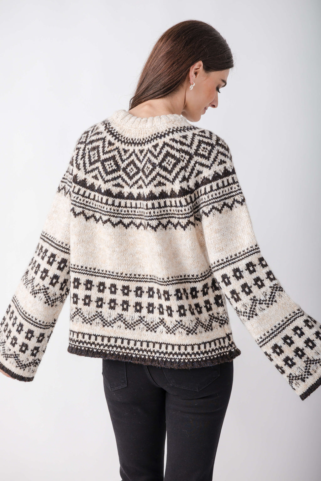 A woman seen from behind wears a cream knit sweater with black geometric Nordic patterns, wide flared sleeves, and black jeans.