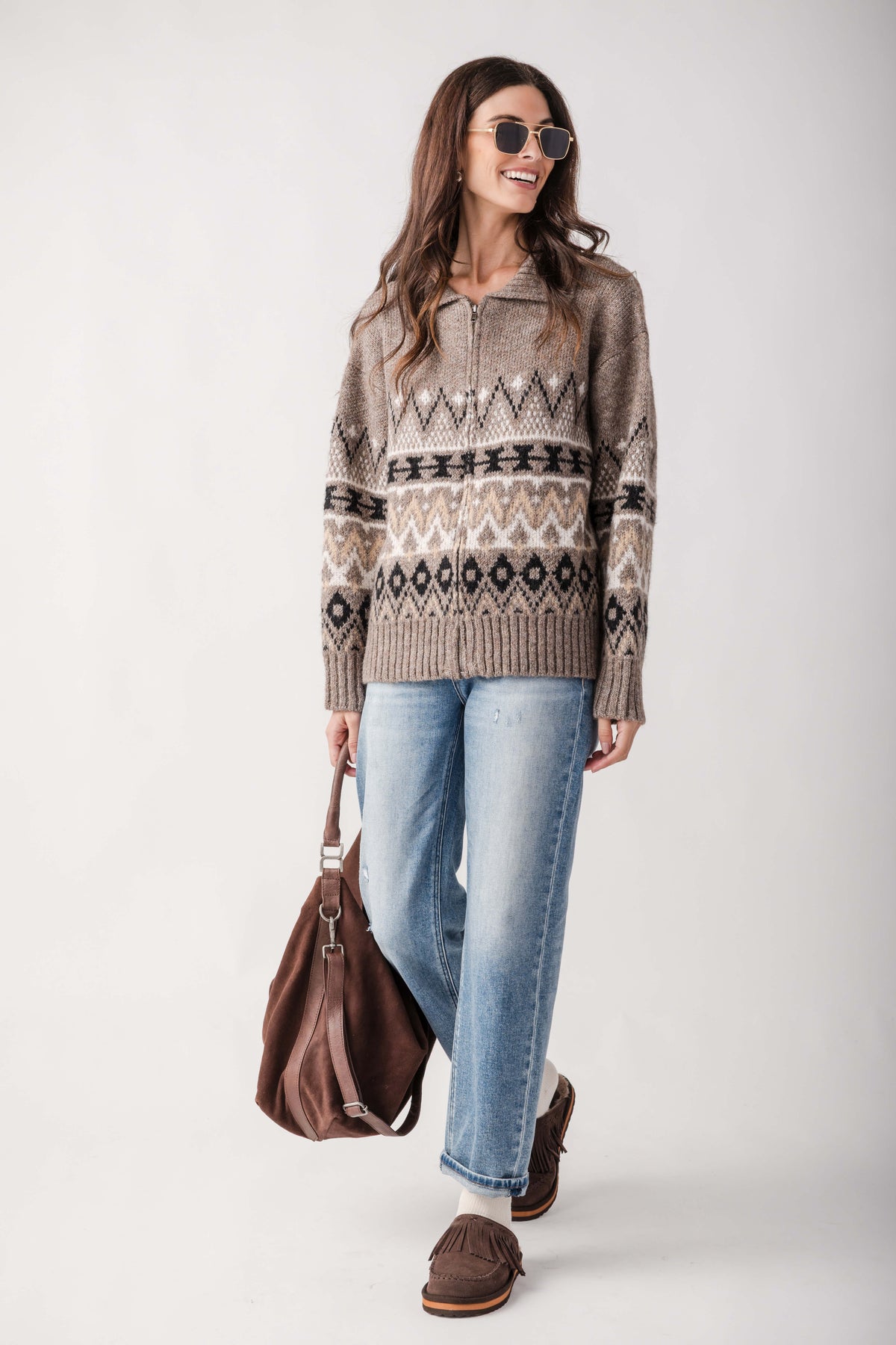 A woman modeling a brown knit sweater with a geometric Fair Isle pattern, paired with light-wash blue jeans, brown loafers, and carrying a brown leather bag against a light gray studio background.