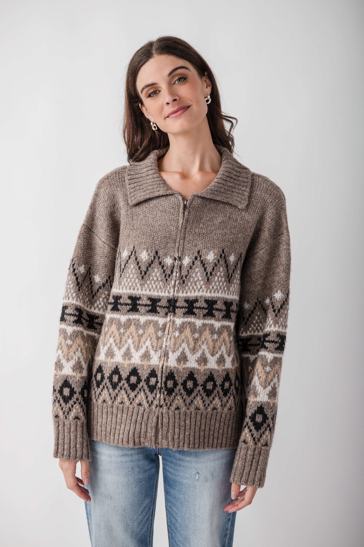 A woman wears a taupe knit zip-up sweater with a ribbed collar and black, cream and tan geometric patterns, paired with light jeans.