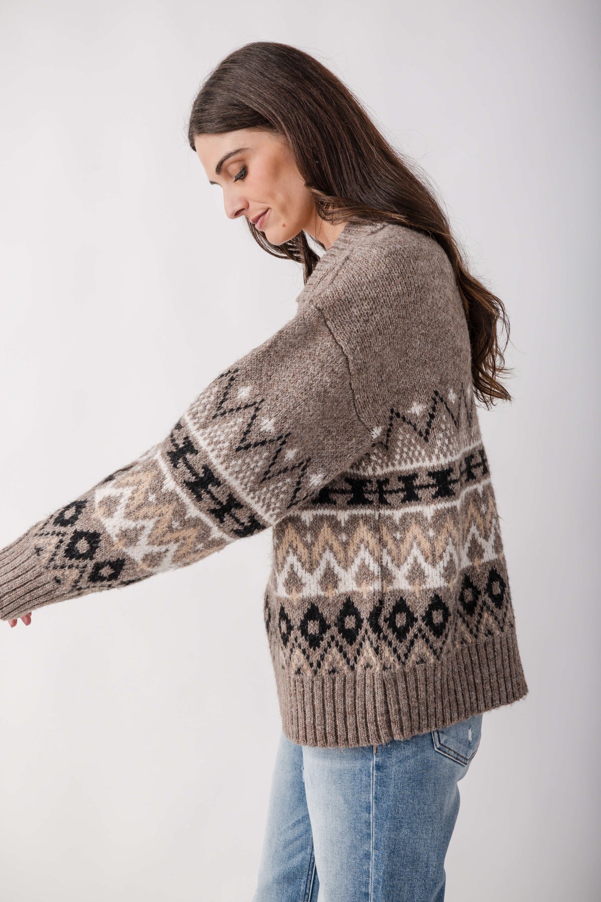 A woman with long brown hair wears a taupe knit sweater with black, cream and tan patterned bands, paired with light blue jeans.