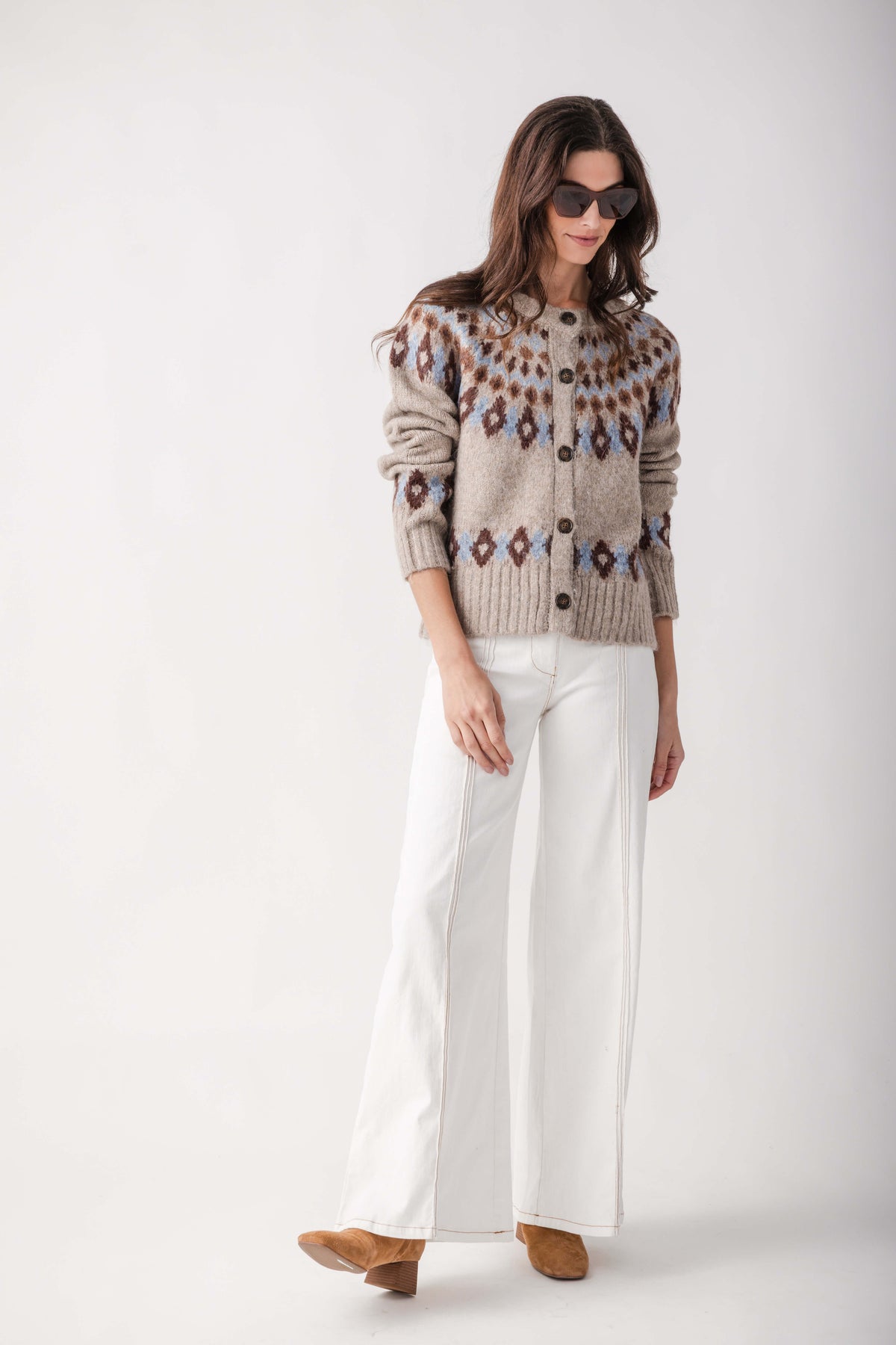 A woman modeling a beige knit cardigan with a brown and blue geometric pattern, paired with white wide-leg pants and brown suede shoes, against a plain white studio background.