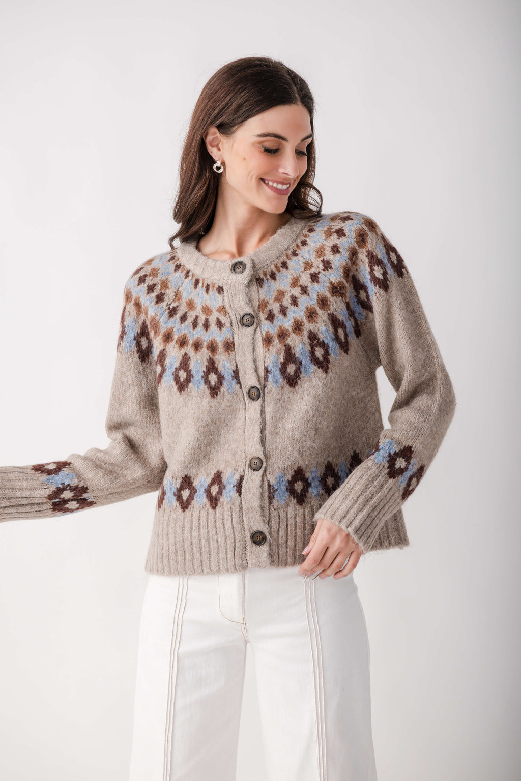 A woman wearing a beige knit cardigan with brown and light blue Fair Isle patterns, paired with white pants, against a plain light gray background.