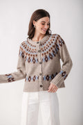 A woman wearing a beige knit cardigan with brown and light blue Fair Isle patterns, paired with white pants, against a plain light gray background.
