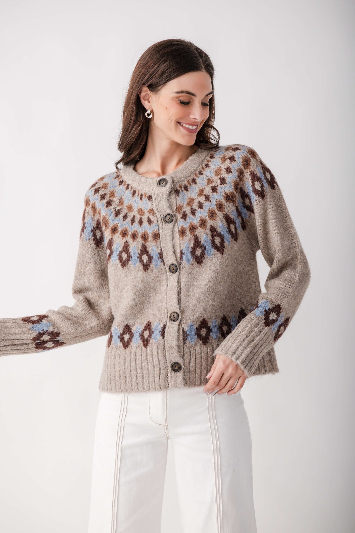 A woman wearing a beige knit cardigan with brown and light blue Fair Isle patterns, paired with white pants, against a plain light gray background.