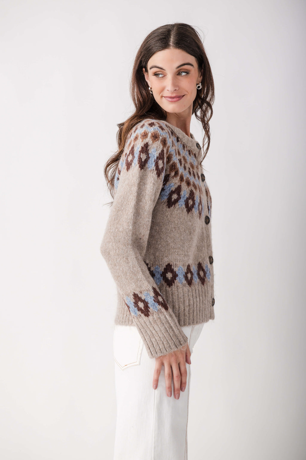 A woman wears a beige button-front knit cardigan with brown and blue Fair Isle patterns, paired with white pants, glancing over her shoulder.