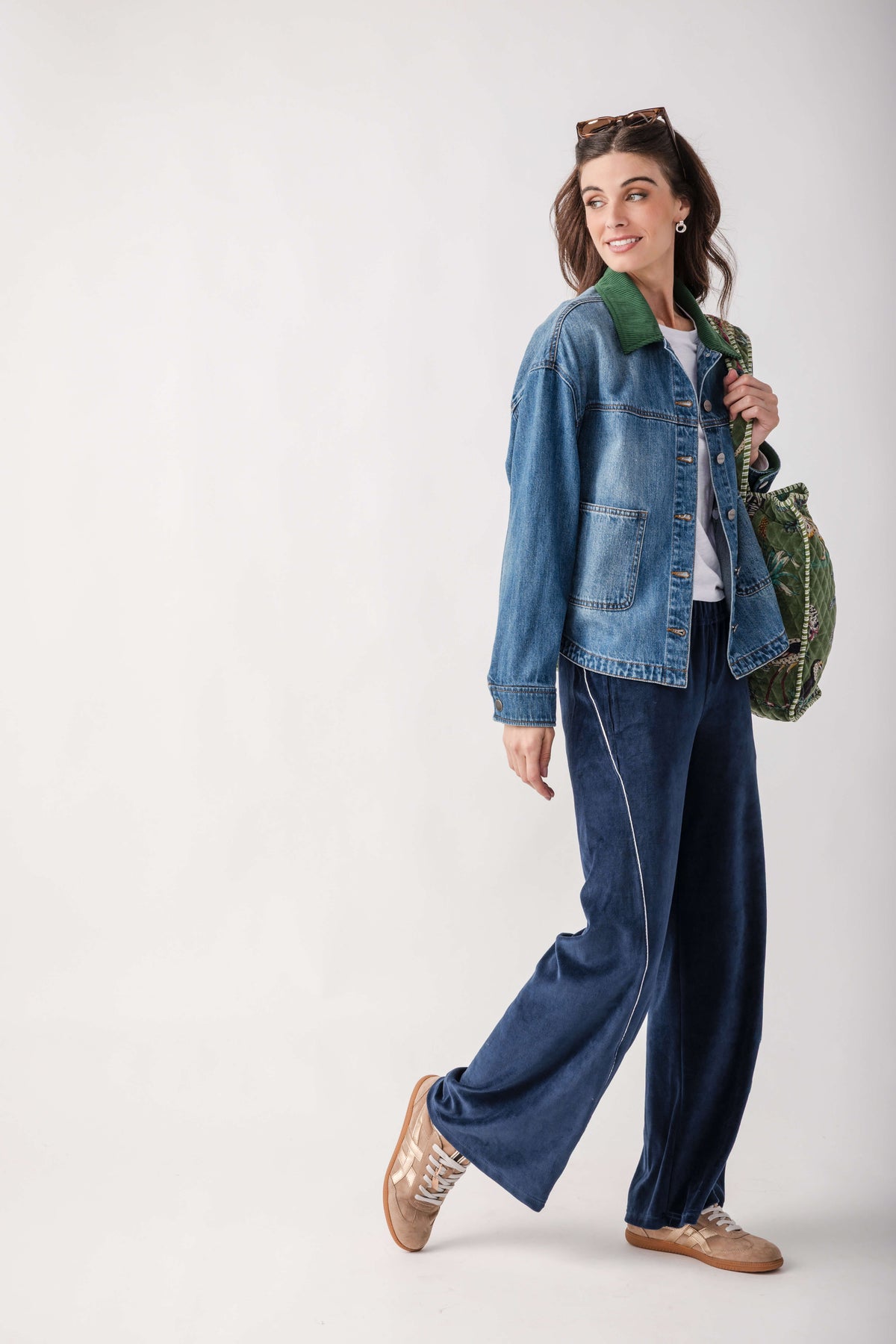 A woman modeling a blue denim jacket with a green collar, paired with dark blue wide-leg trousers, metallic gold sneakers, and carrying a green patterned shoulder bag against a plain light gray studio background.