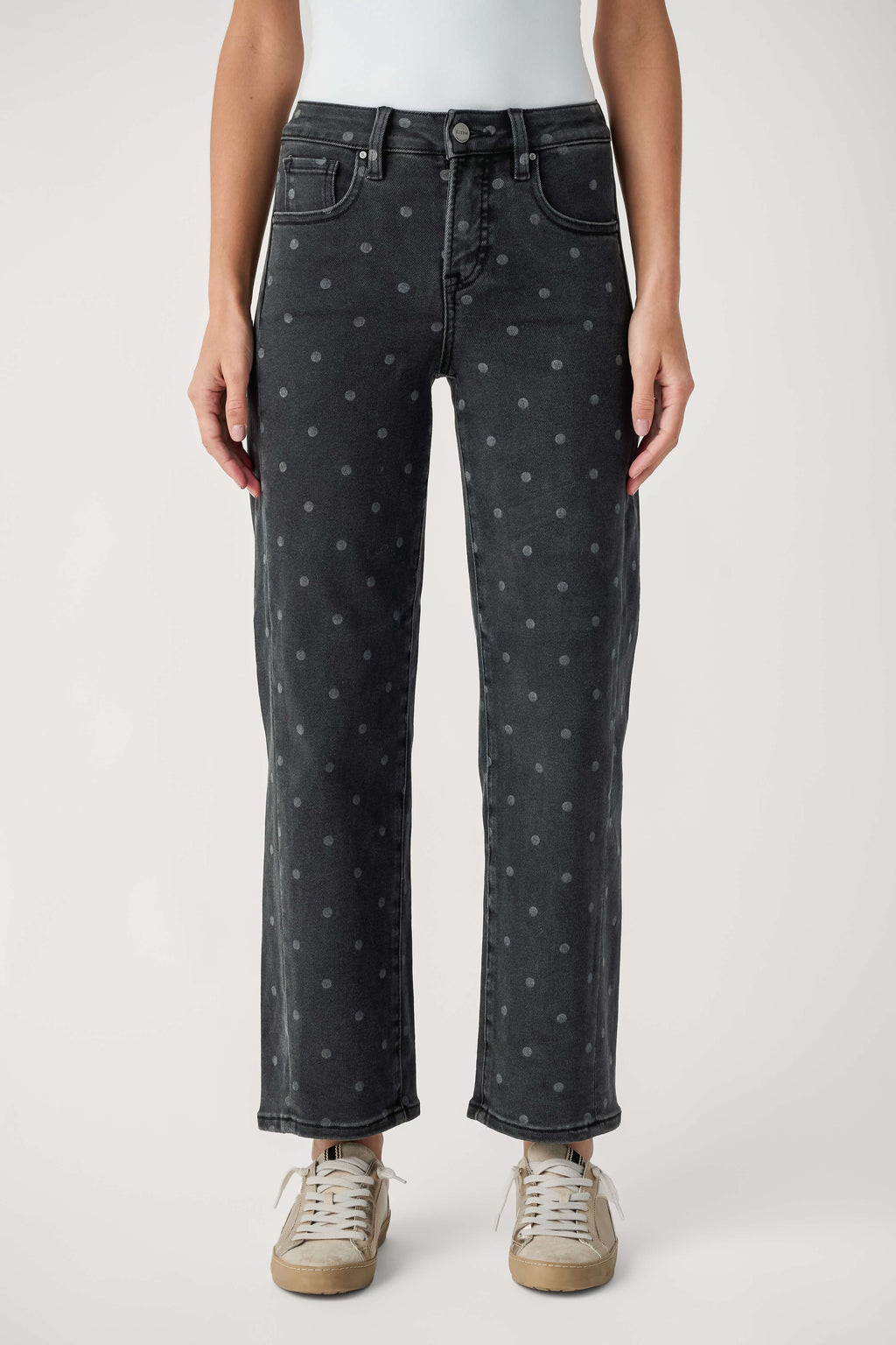 A model from the waist down wearing dark gray washed jeans with a small light gray polka dot pattern, paired with beige sneakers, against a light gray studio background.