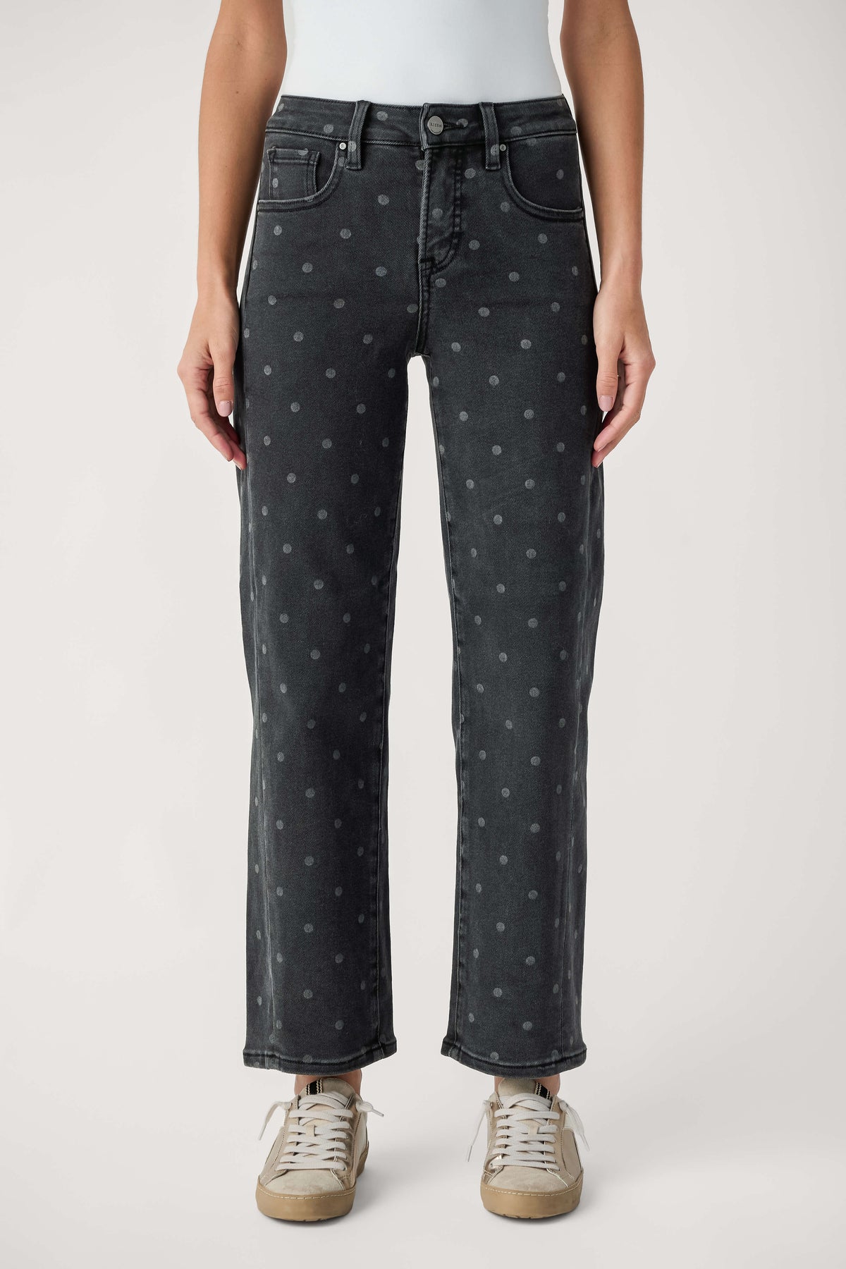 A model from the waist down wearing dark gray washed jeans with a small light gray polka dot pattern, paired with beige sneakers, against a light gray studio background.