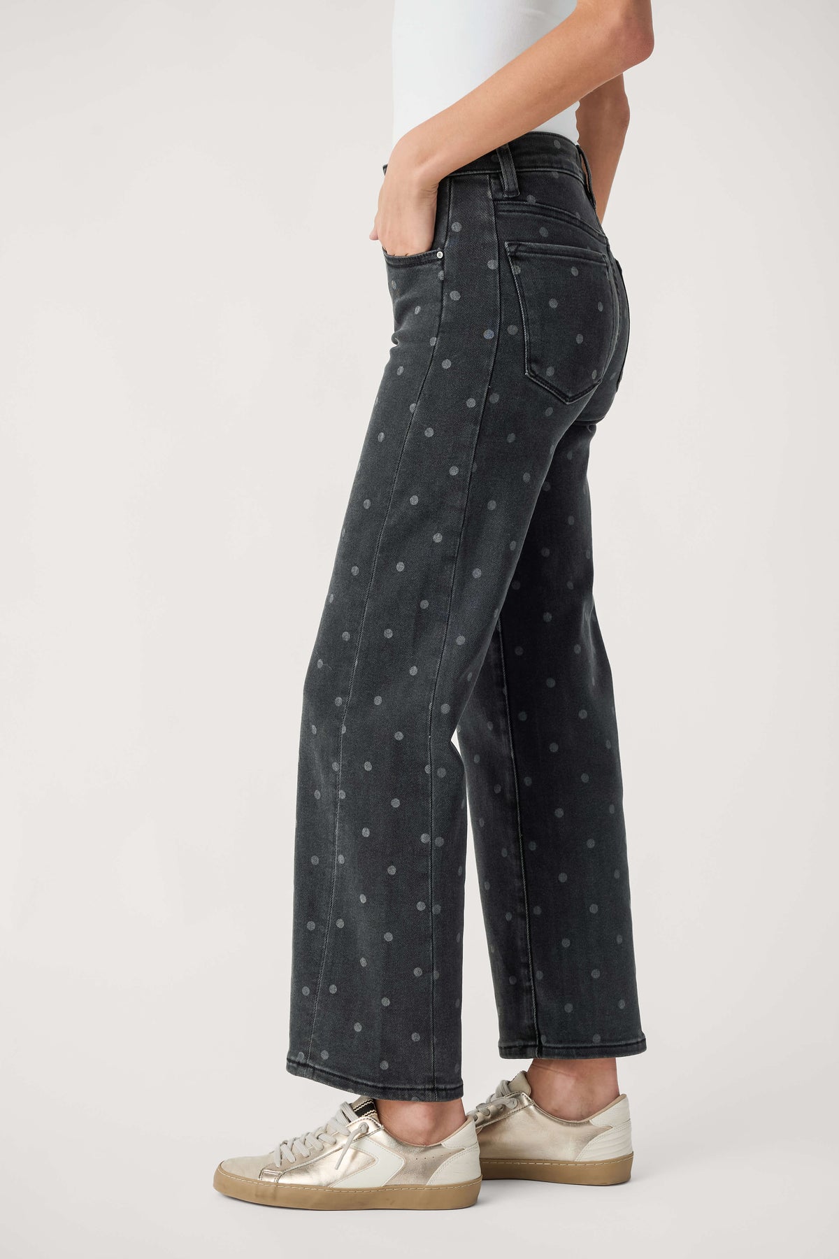 A person wears black high-rise ankle jeans with small gray polka dots, a white top, and metallic gold sneakers.