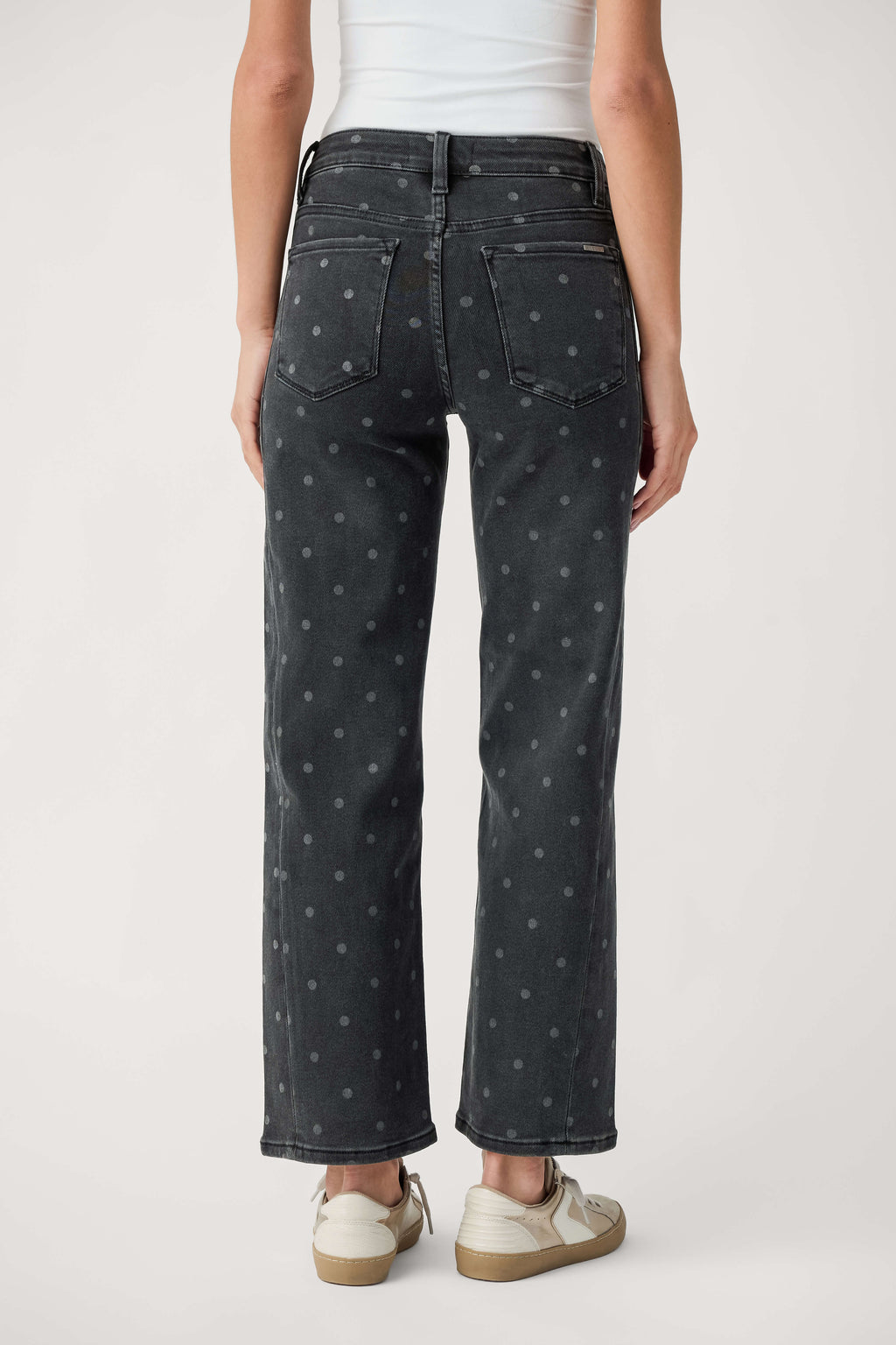Rear view of a person wearing faded black ankle-length jeans with small gray polka dots, a white top, and cream sneakers.