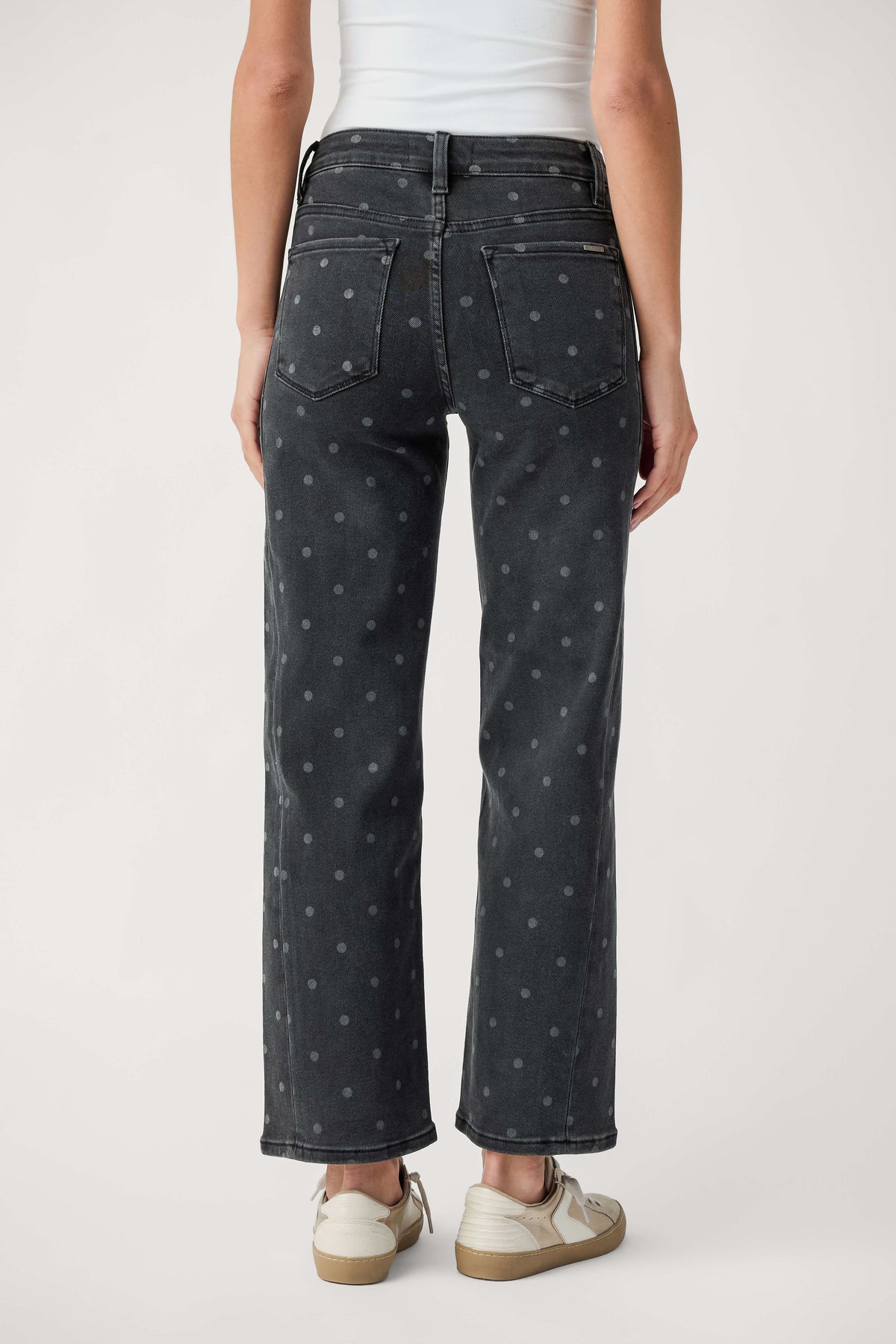 Rear view of a person wearing faded black ankle-length jeans with small gray polka dots, a white top, and cream sneakers.