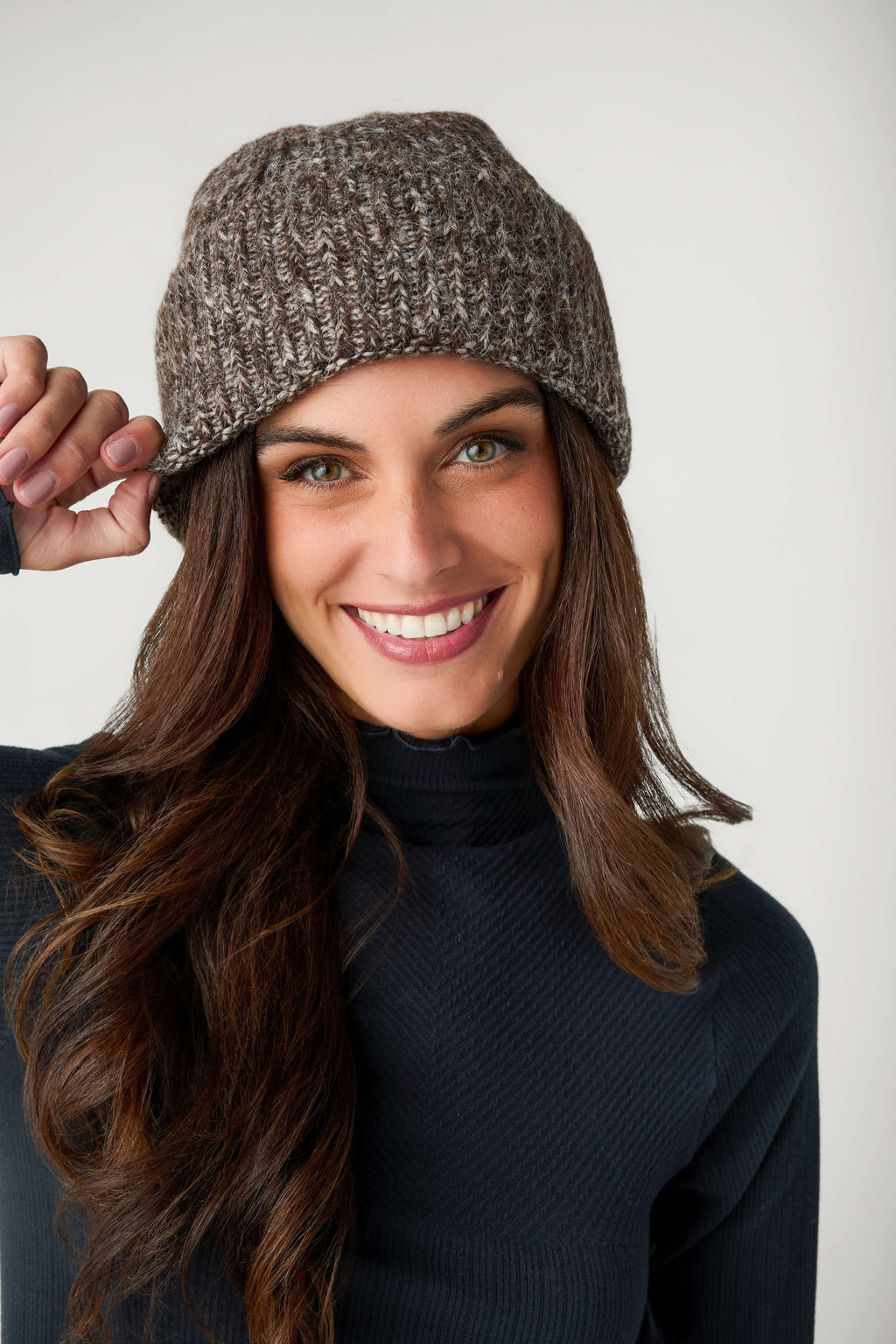 A smiling woman with long brown hair wears a brown and cream marled knit beanie and a black turtleneck.