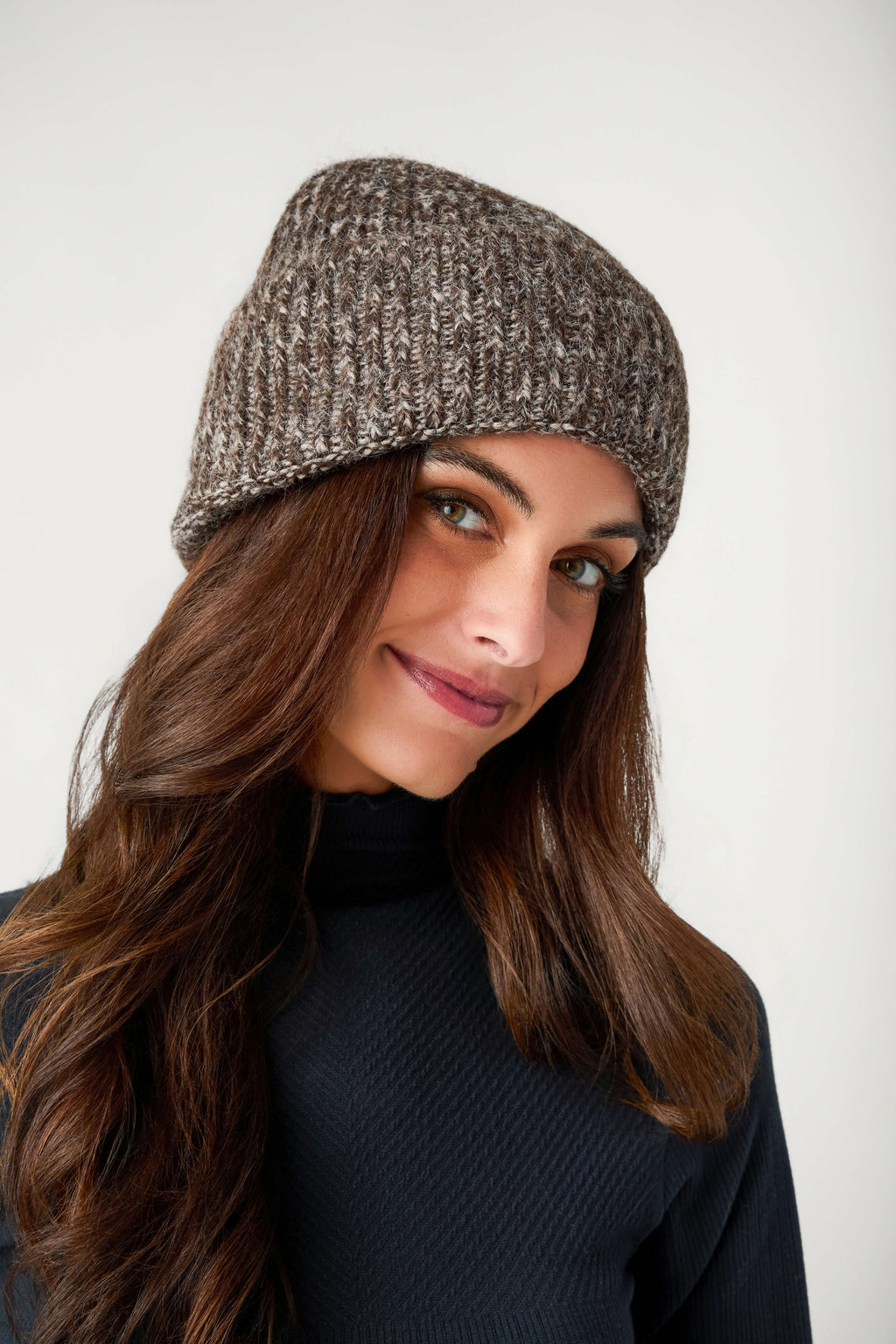 A smiling woman with long brown hair wears a brown and cream marled knit beanie with a black turtleneck.