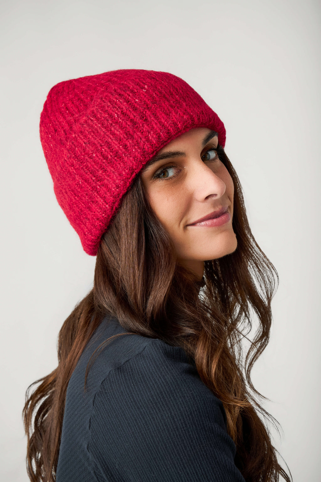 A woman with long brown hair wears a red ribbed knit beanie and a dark navy top against a plain background.