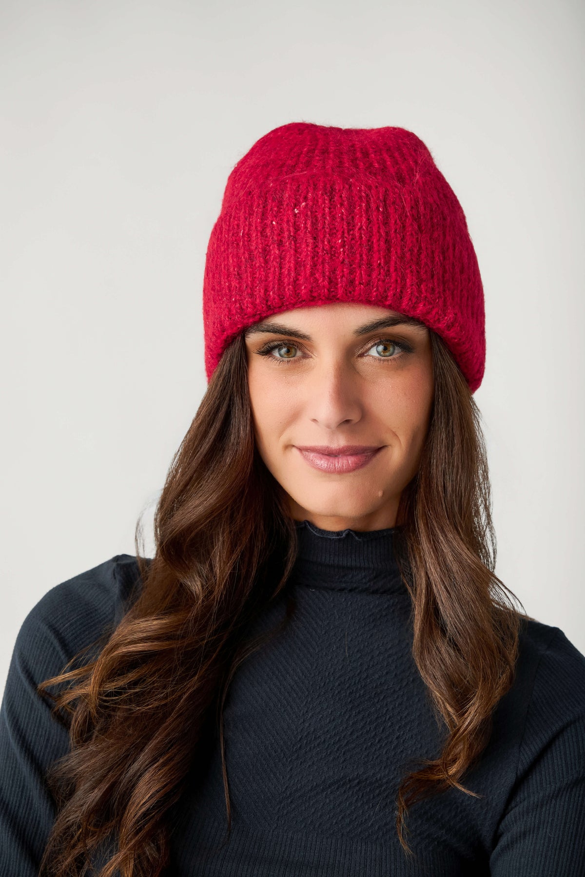 A woman with long wavy brown hair wears a red ribbed knit beanie and a dark navy turtleneck top.