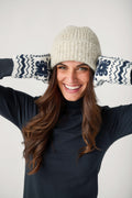 A smiling woman with long brown hair wears a cream knit beanie, navy turtleneck, and navy-and-white patterned mittens raised beside her head.