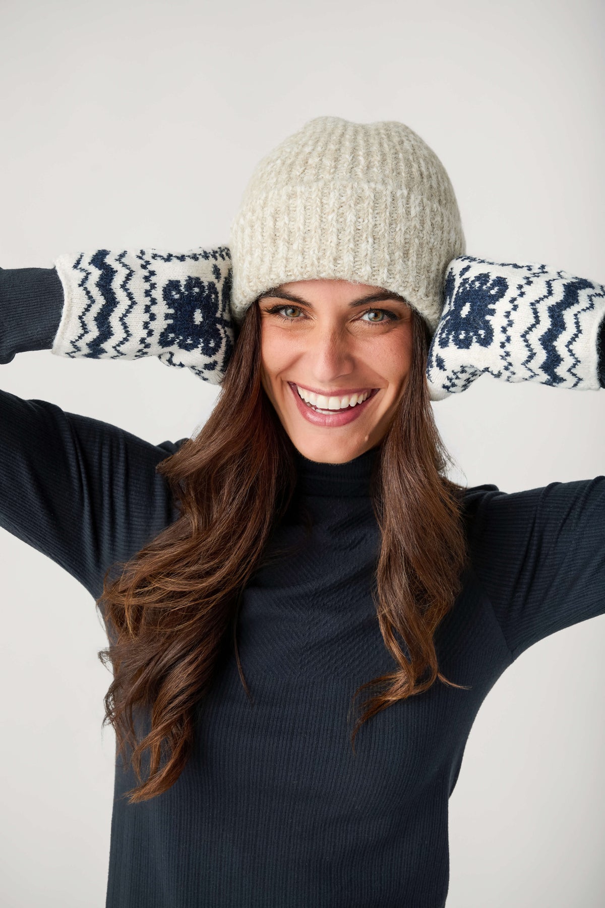 A smiling woman with long brown hair wears a cream knit beanie, navy turtleneck, and navy-and-white patterned mittens raised beside her head.