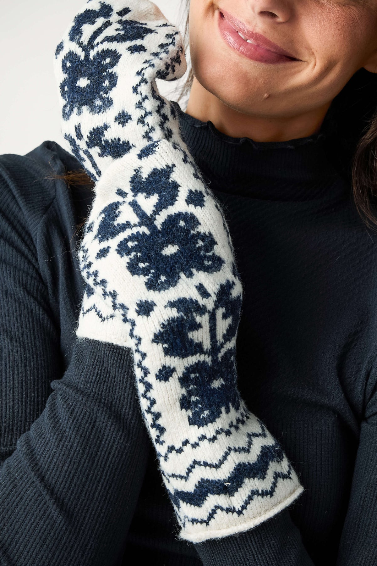 A smiling woman in a dark navy ribbed top wears cream knit mittens patterned with navy floral and zigzag designs.
