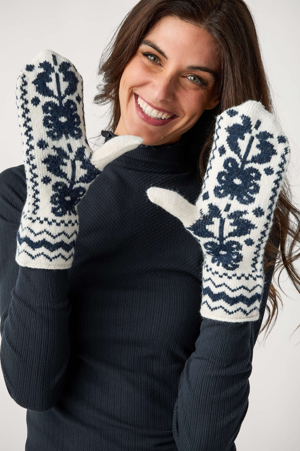 A smiling woman with long brown hair wears cream knit mittens with navy floral and zigzag patterns over a dark ribbed top.