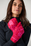 A woman in a black ribbed turtleneck holds up red knit mittens with a pink floral pattern in front of her chest.