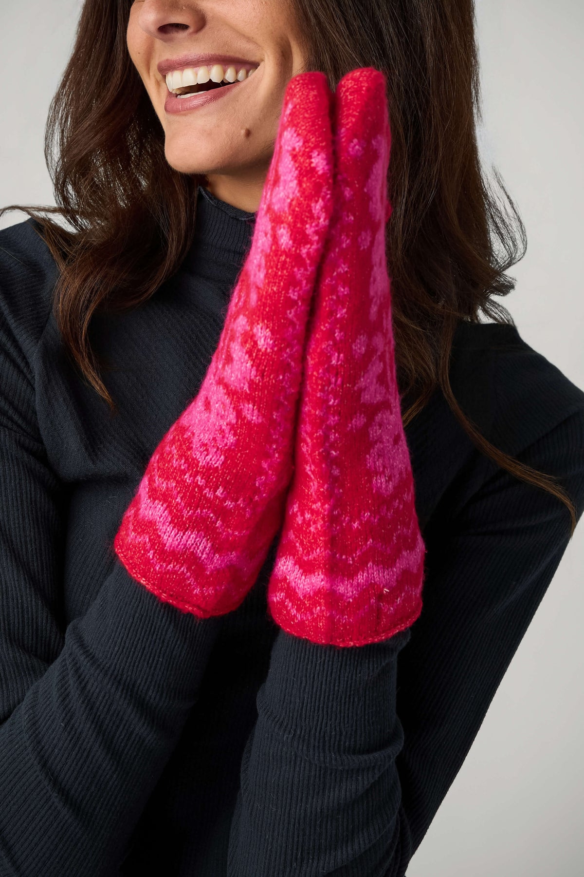 A smiling woman in a black ribbed top holds her hands together, wearing red mittens with a pink folk-style knit pattern.