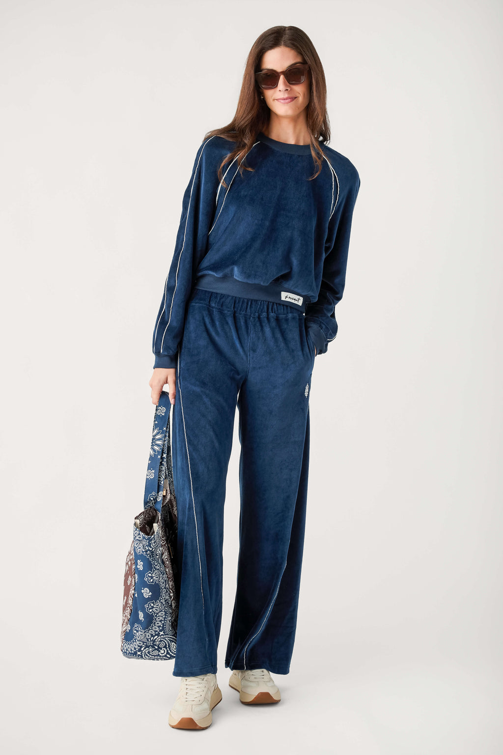 A woman wears navy blue velour wide-leg pants with white piping and a matching sweatshirt, holding a paisley tote bag.