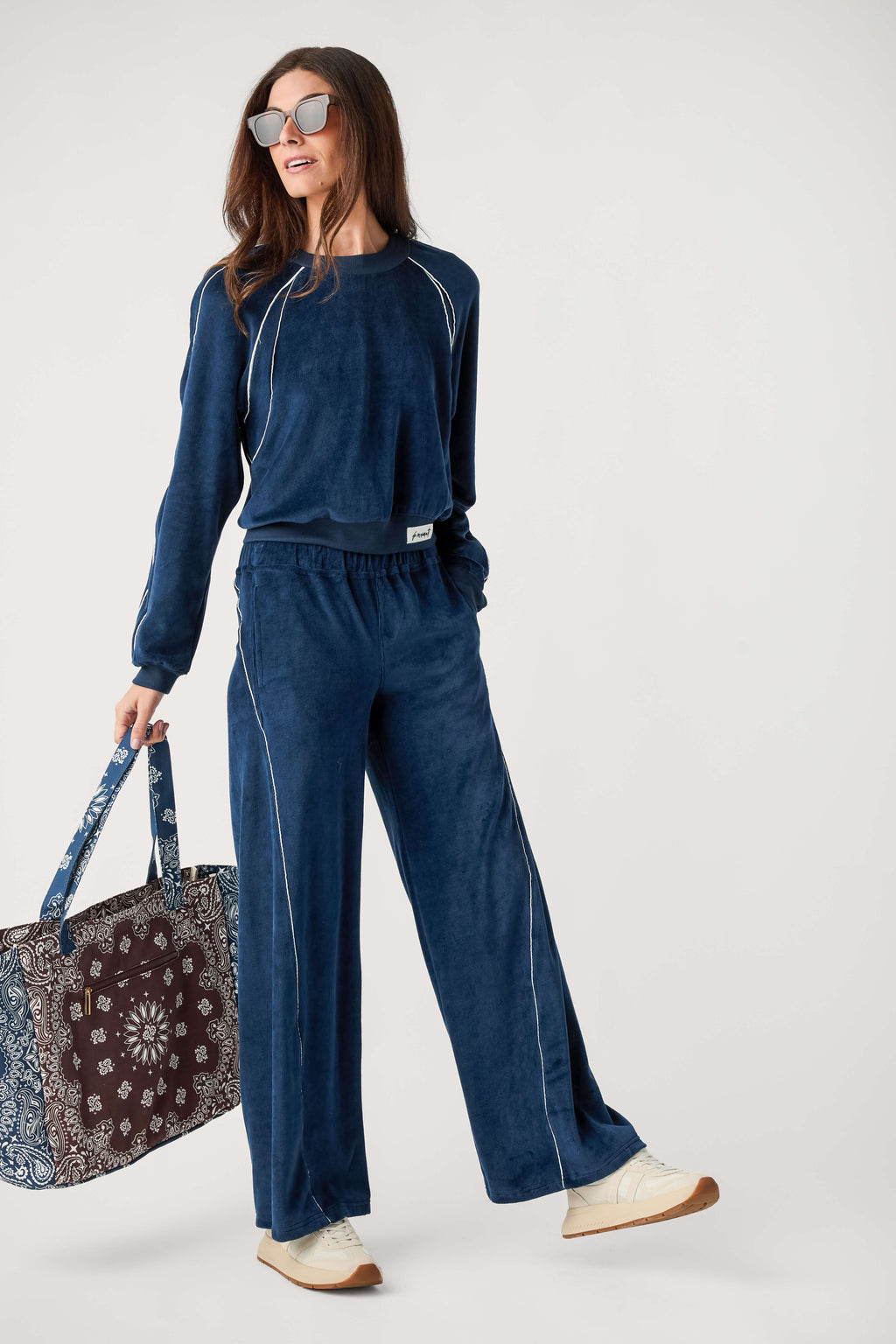 A woman wears a navy velour pullover with white piping and matching wide-leg pants, carrying a paisley tote bag.
