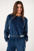 A woman with long brown hair wears a navy velour crewneck pullover with white piping and matching velour pants.