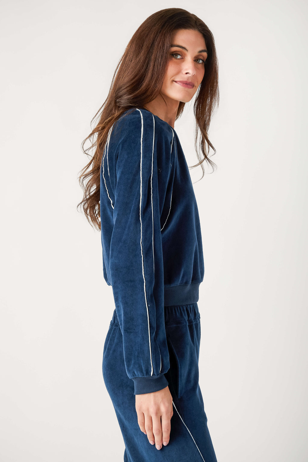 A woman with long brown hair wears a navy blue velour pullover with white piping along the sleeves and matching pants.