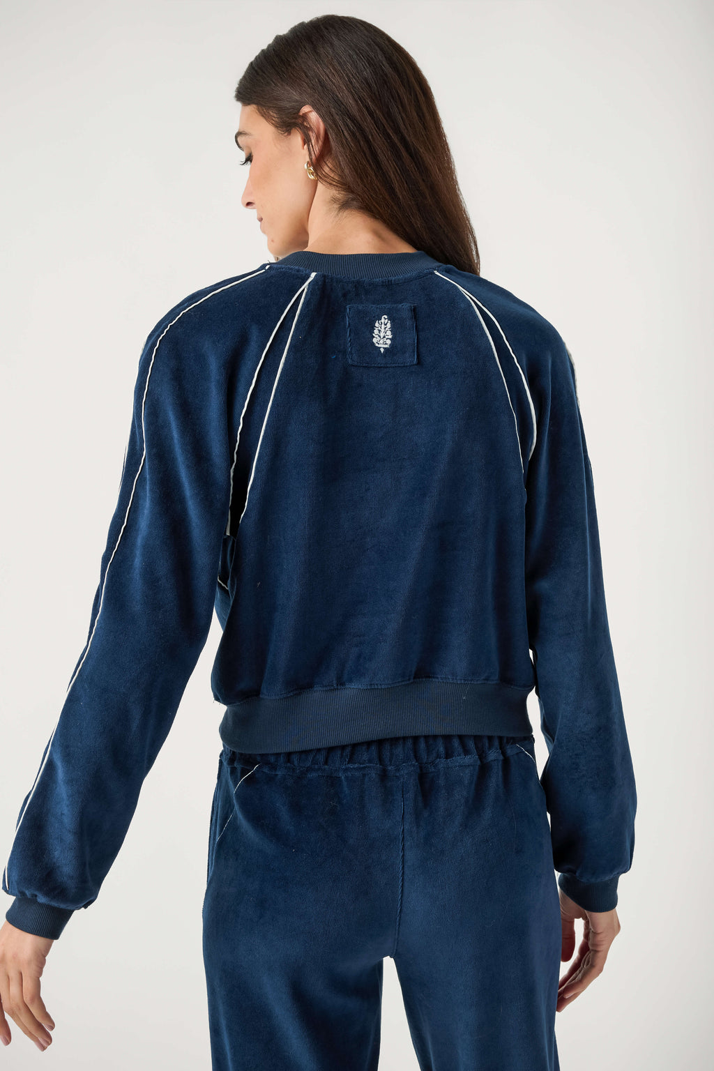 Back view of a woman wearing a navy velour cropped jacket with white piping, ribbed trim, and matching navy pants.