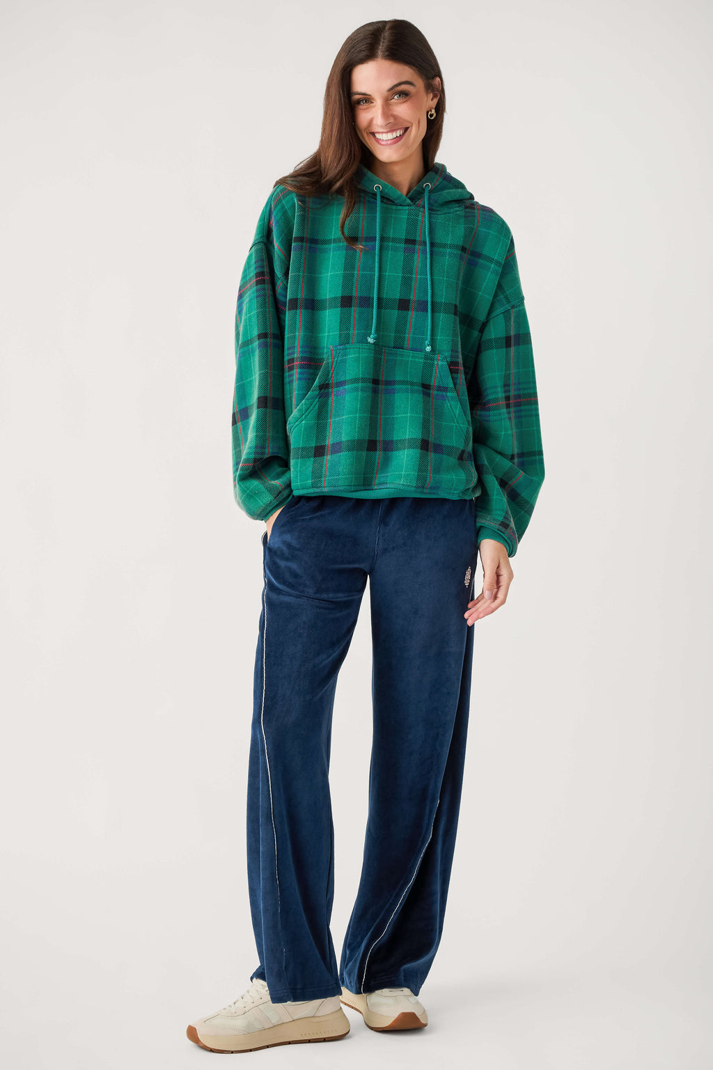 A smiling woman wears a green plaid hooded sweatshirt with a front pocket, navy velour wide-leg pants, and cream sneakers.