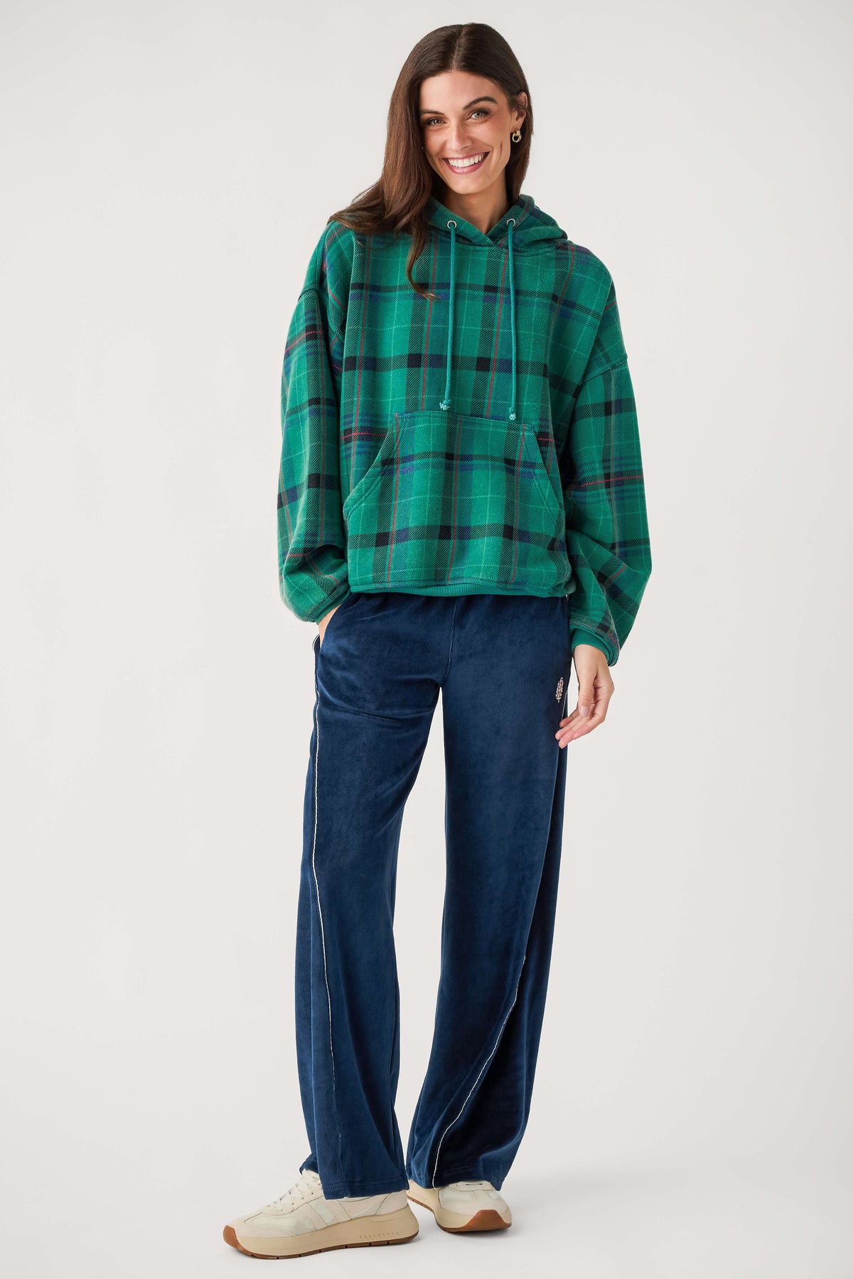 A smiling woman wears a green plaid hooded sweatshirt with a front pocket, navy velour wide-leg pants, and cream sneakers.