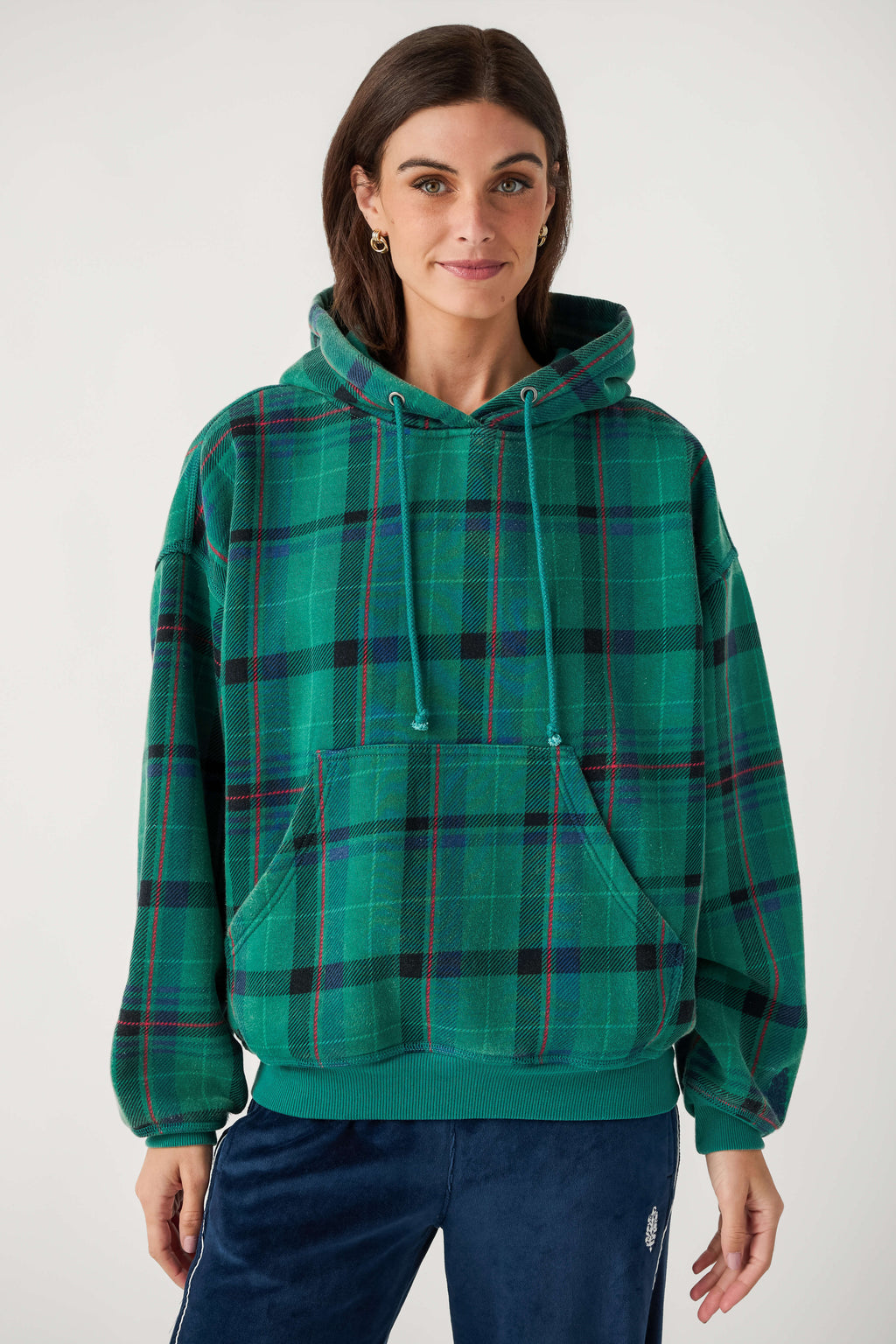 A woman wears a green plaid hooded sweatshirt with red and black stripes, a front pouch pocket, and navy velour pants.